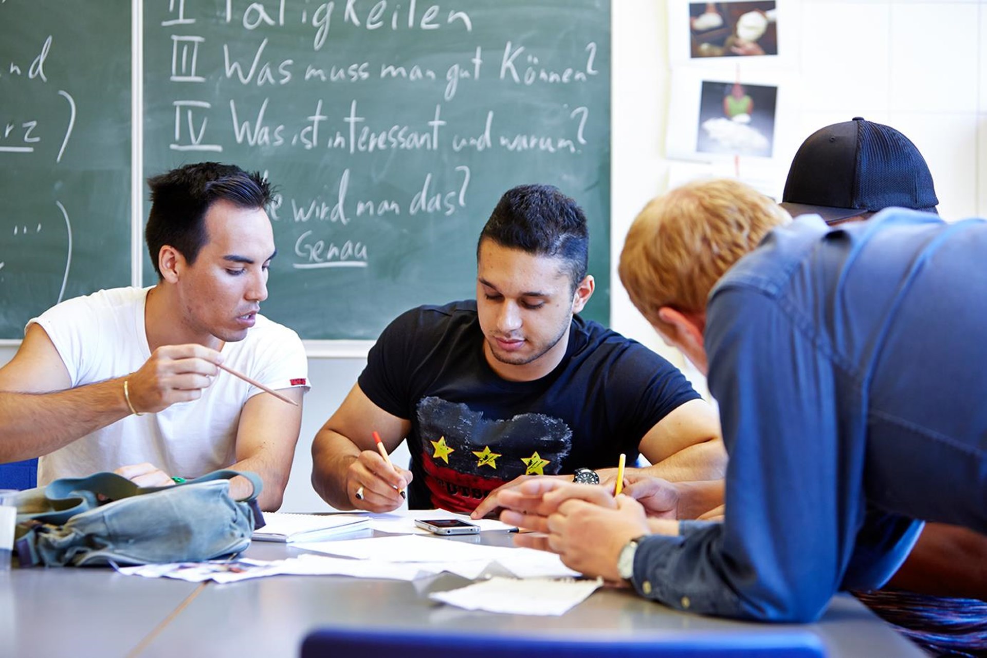 Drei junge Männer sitzen an einem Tisch, im Hintergrund eine Tafel.